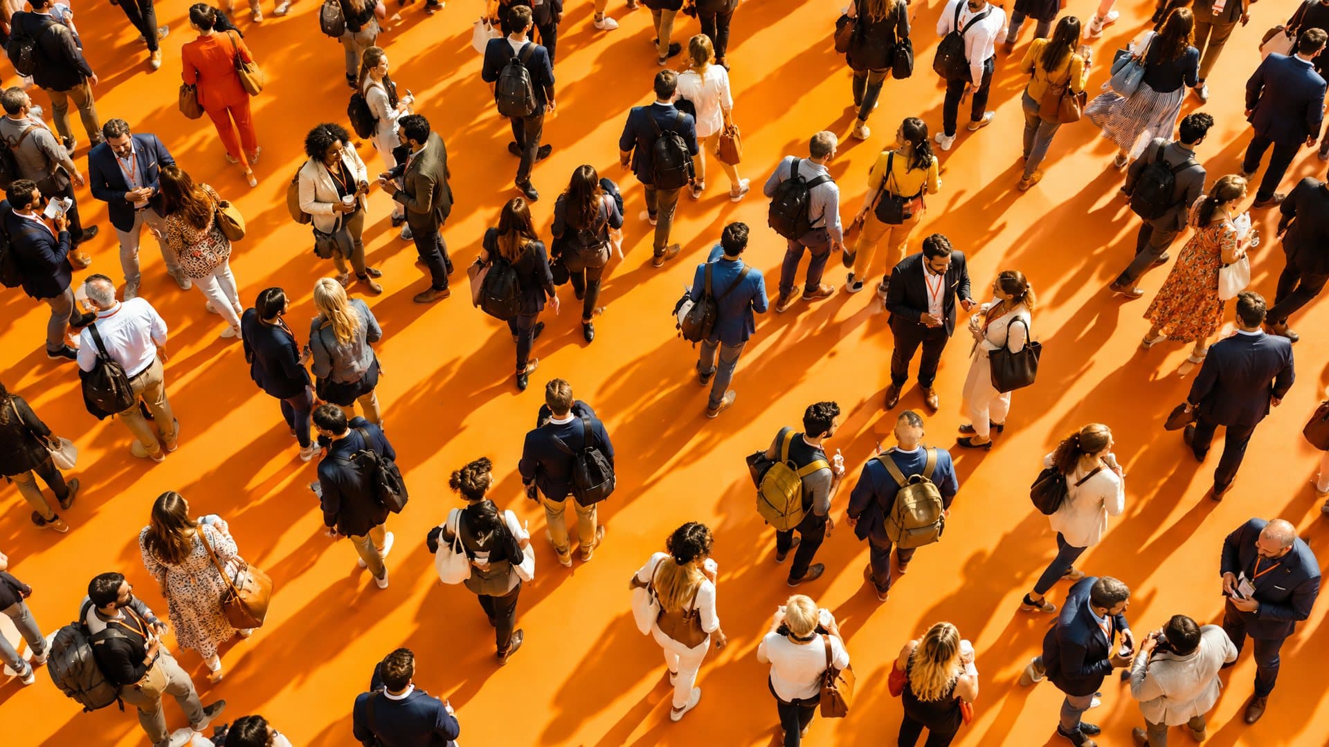 A crowd of conference attendees seen from directly above, crossing a bright orange exhibition floor