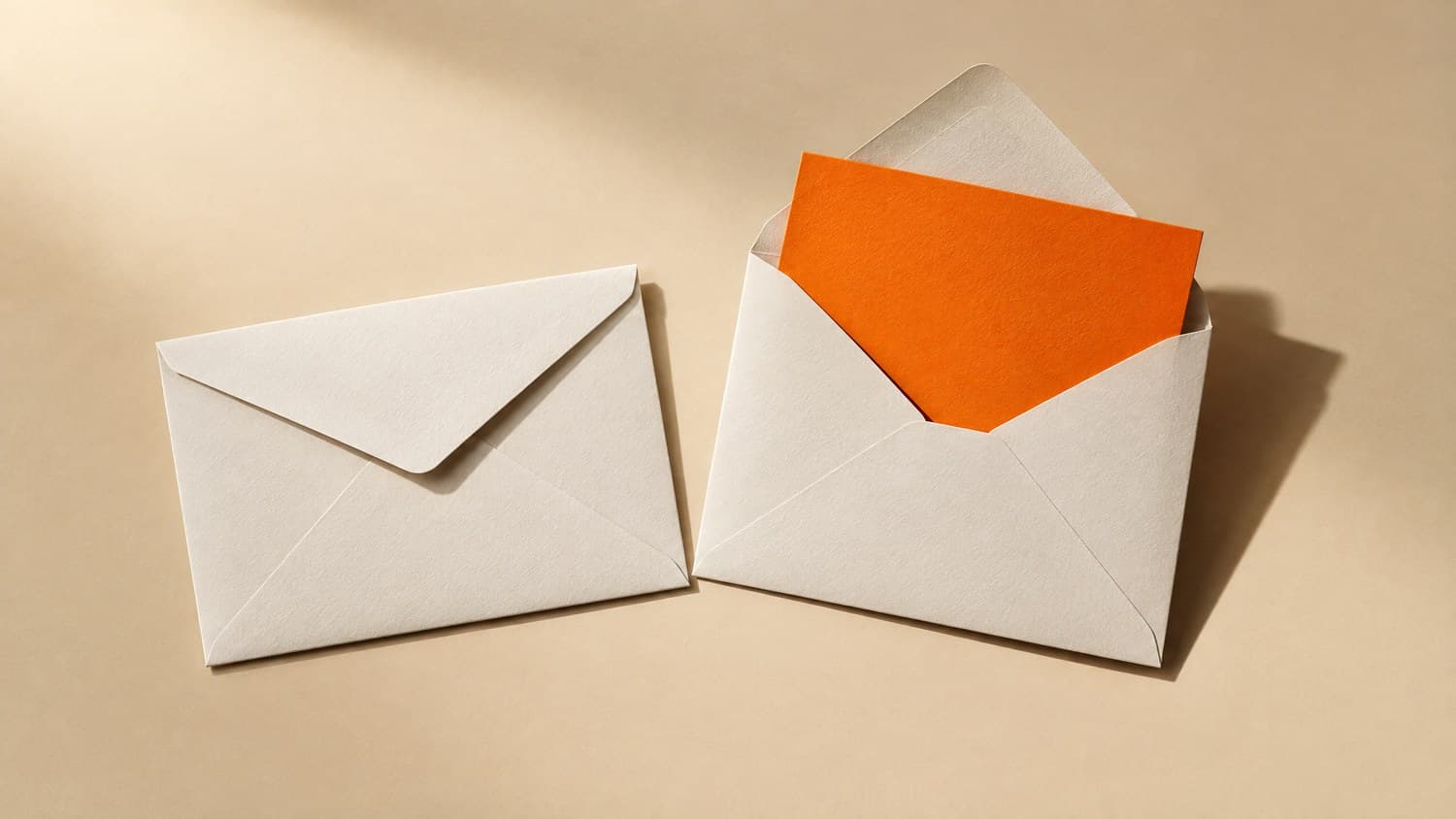A sealed white envelope beside an opened one with a bright orange card drawn part-way out of it